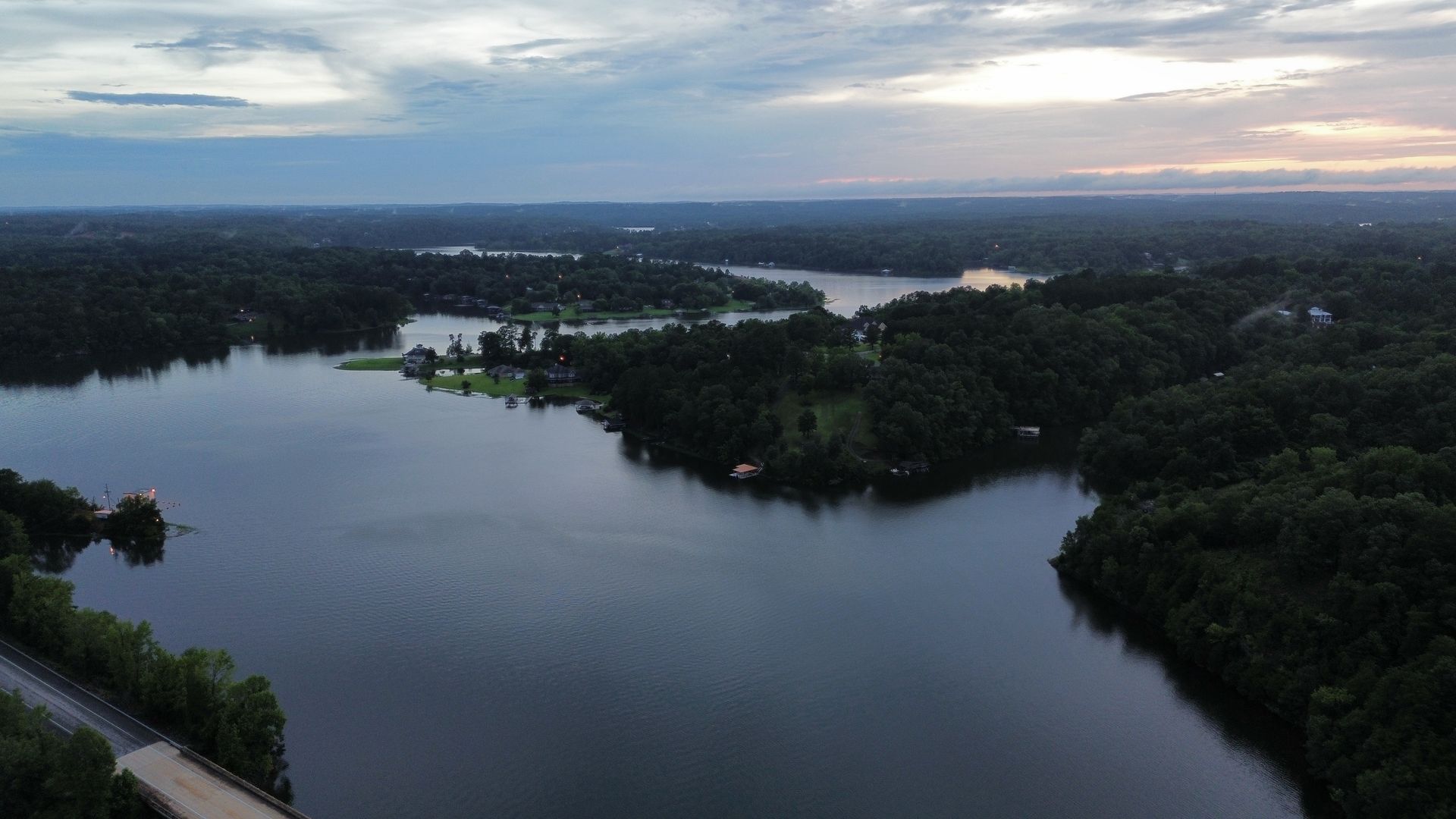 Aerial view of a lake surrounded by lush green trees under a cloudy sky at dusk.