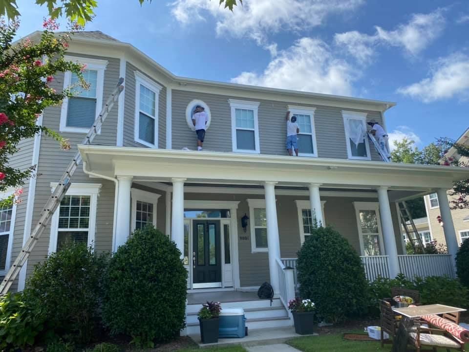 House being painted, workers on roof, gray siding, white trim, blue sky, ladder.