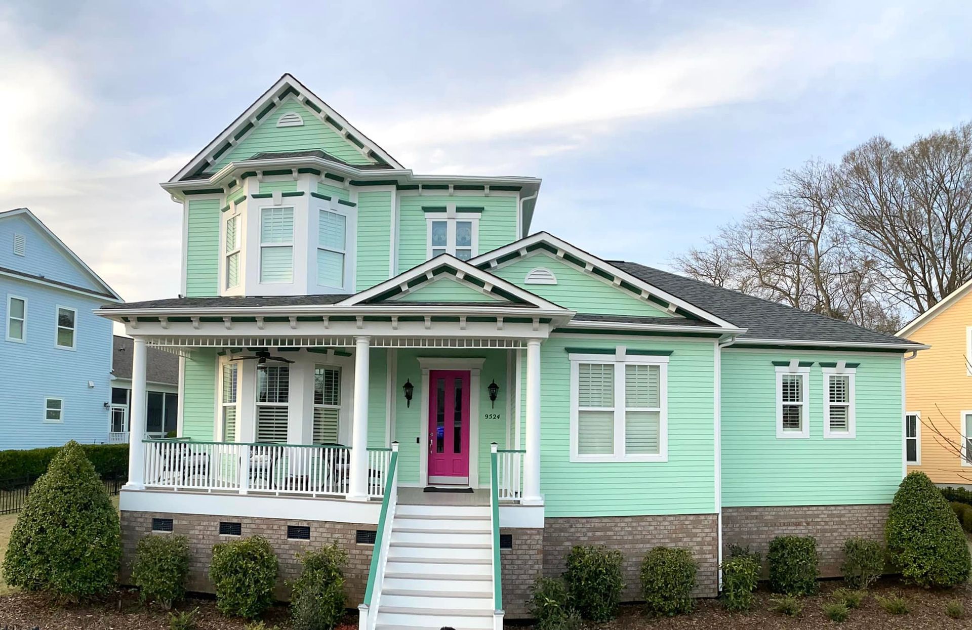 Two-story teal house with a pink door and white trim, porch, and steps; clear sky.