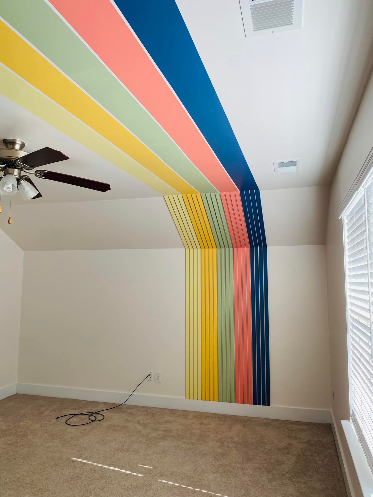 Colorful rainbow stripes painted on wall and ceiling in a room with beige carpet.