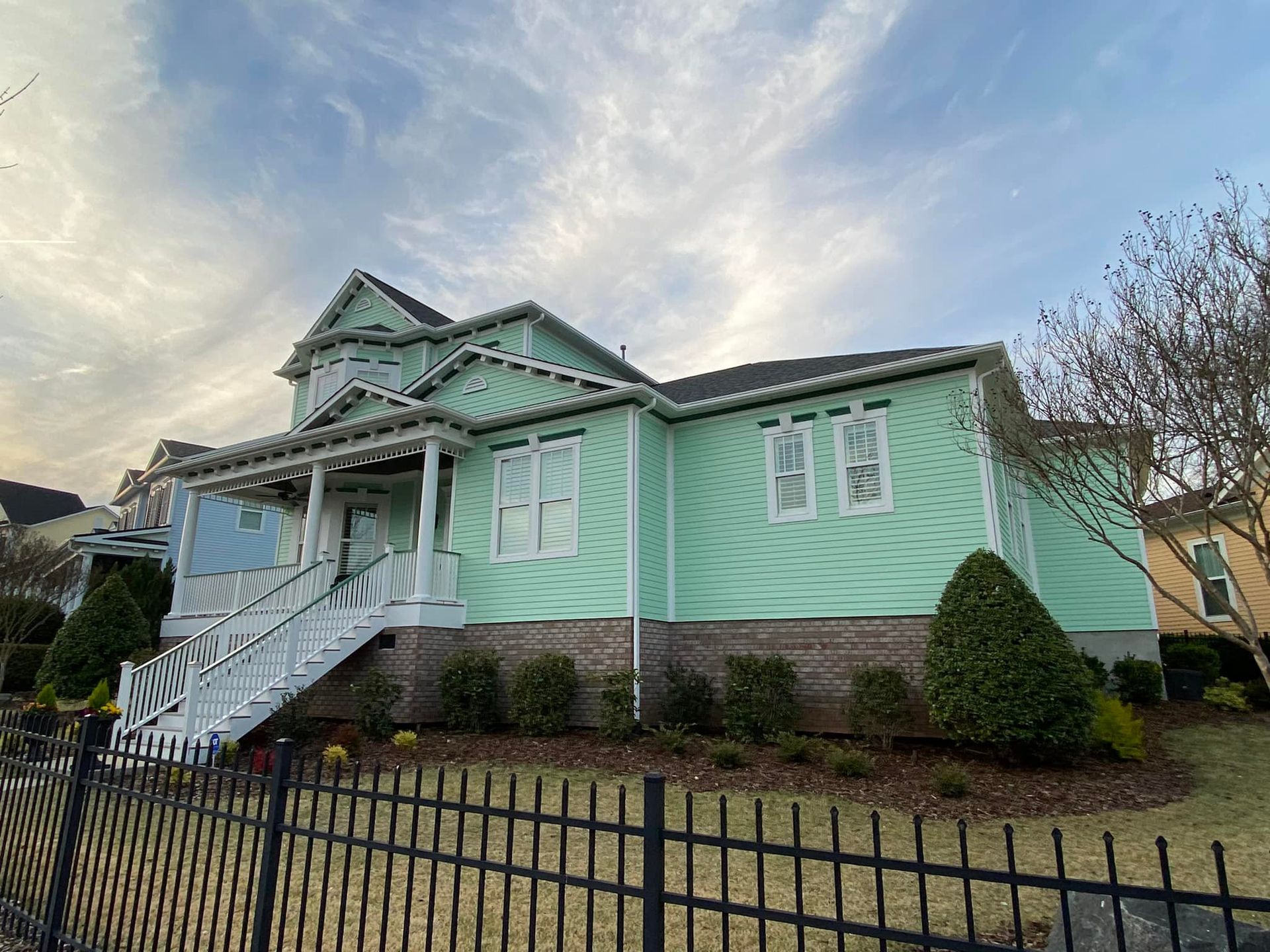 A two-story light green house with white trim, a porch, and a black fence under a cloudy sky.