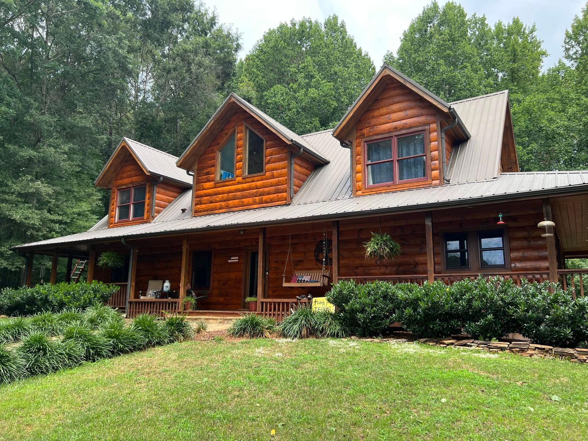 Log cabin with a covered porch and dormers, set in a grassy yard with surrounding trees.