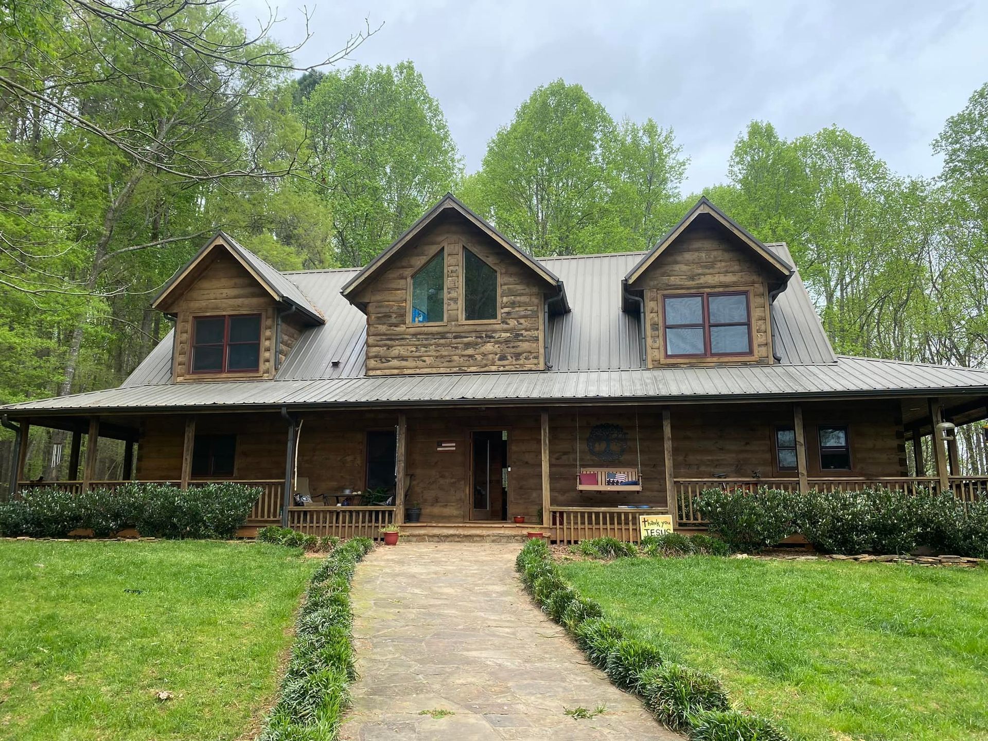 Log cabin home with porch, pathway, and dormers, surrounded by trees and green grass.