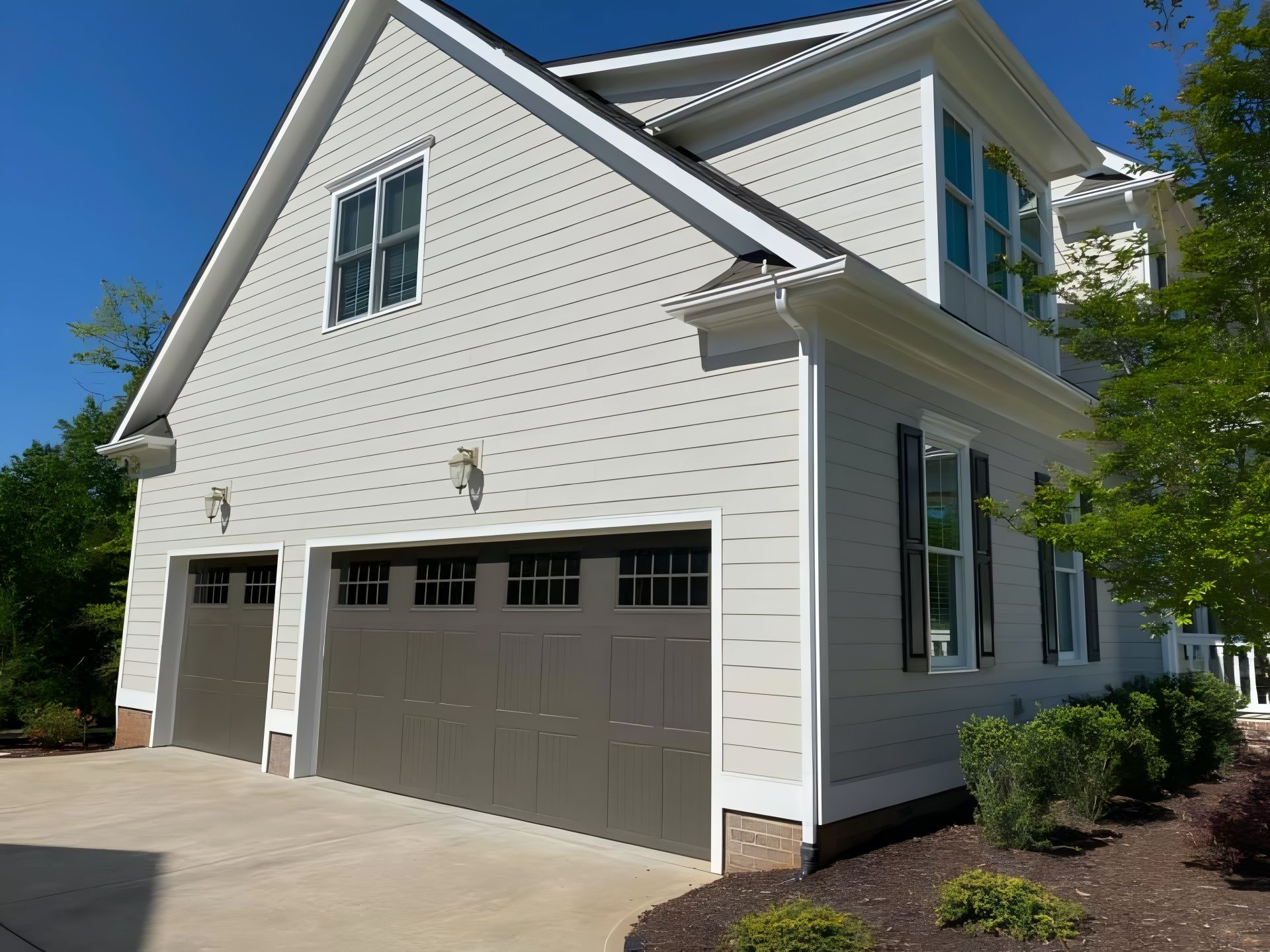 Two-car garage with tan doors and light gray siding, connected to a house with black shutters, on a sunny day.