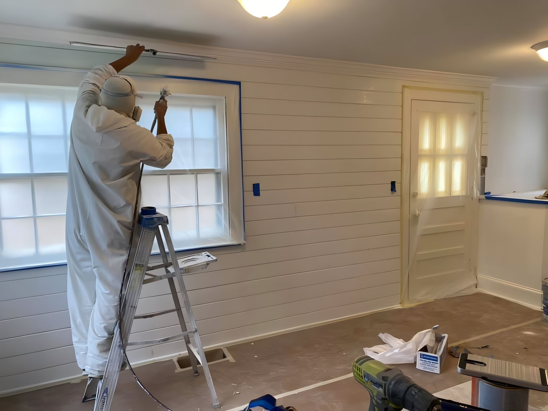 Person in a protective suit sprays white paint on a shiplap wall inside a room.