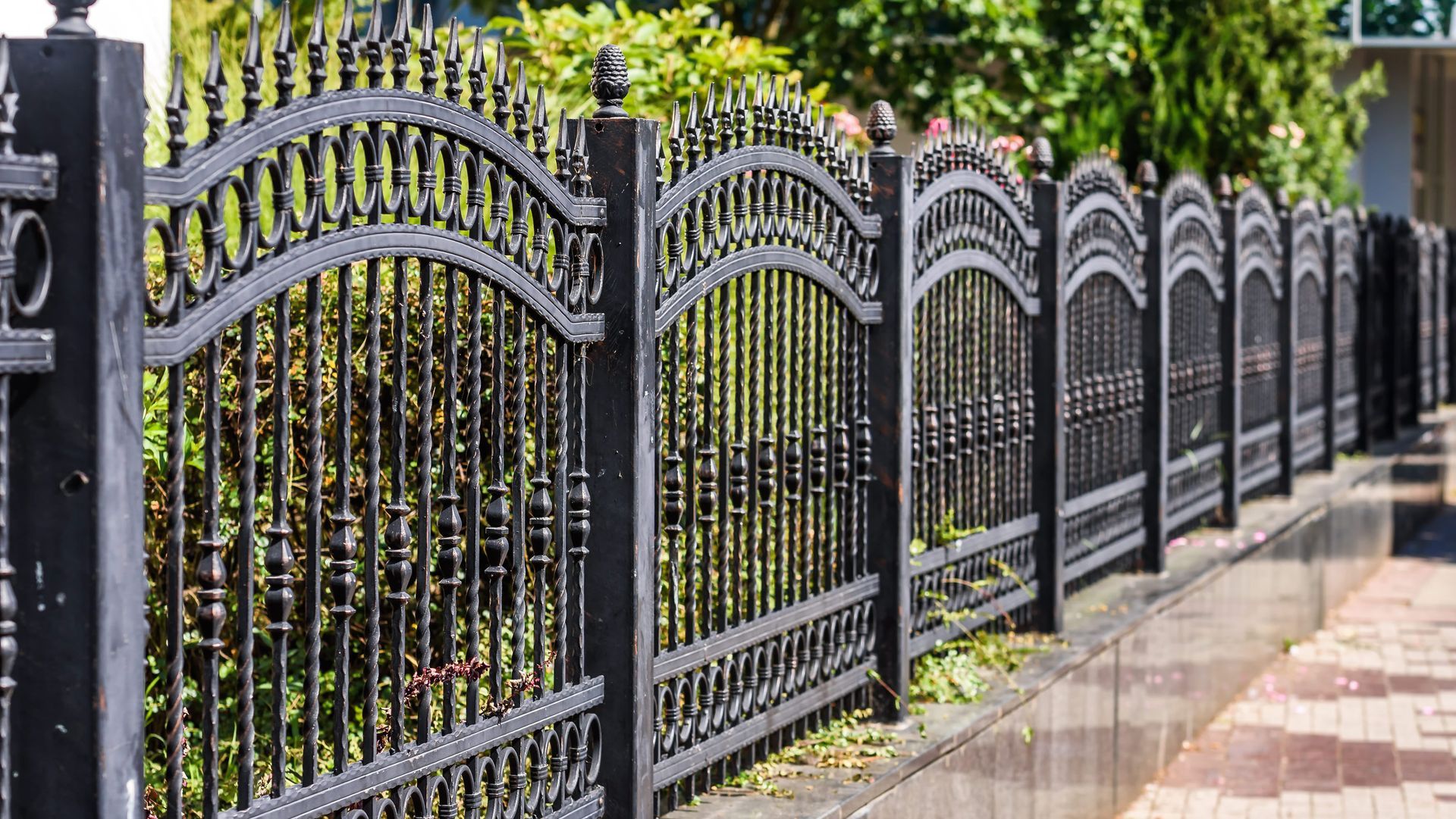 Part of a steel fence that goes around a property with bushes behind it.