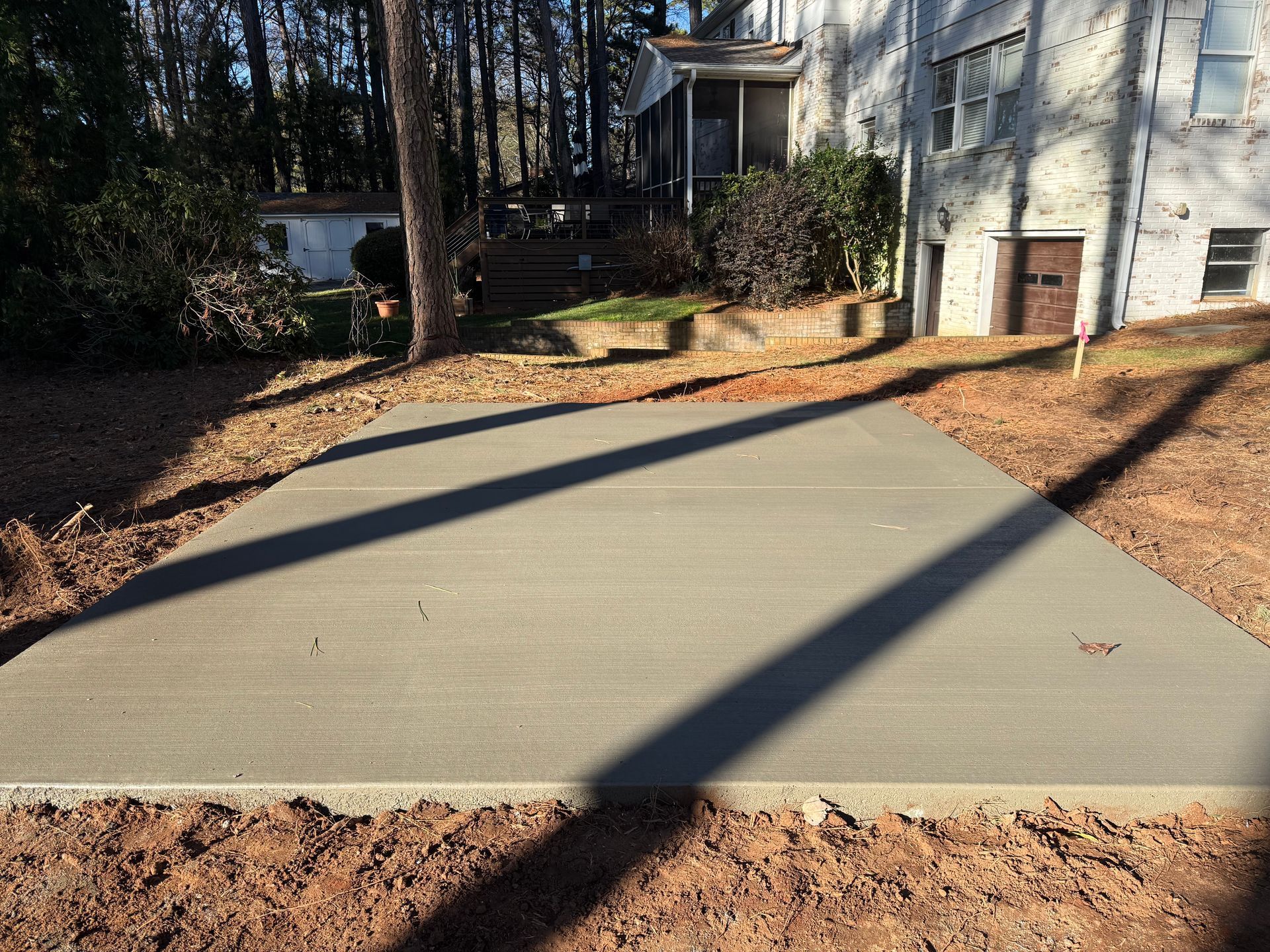 A new rectangular concrete slab sits in a backyard near a house, surrounded by dirt and bare trees.