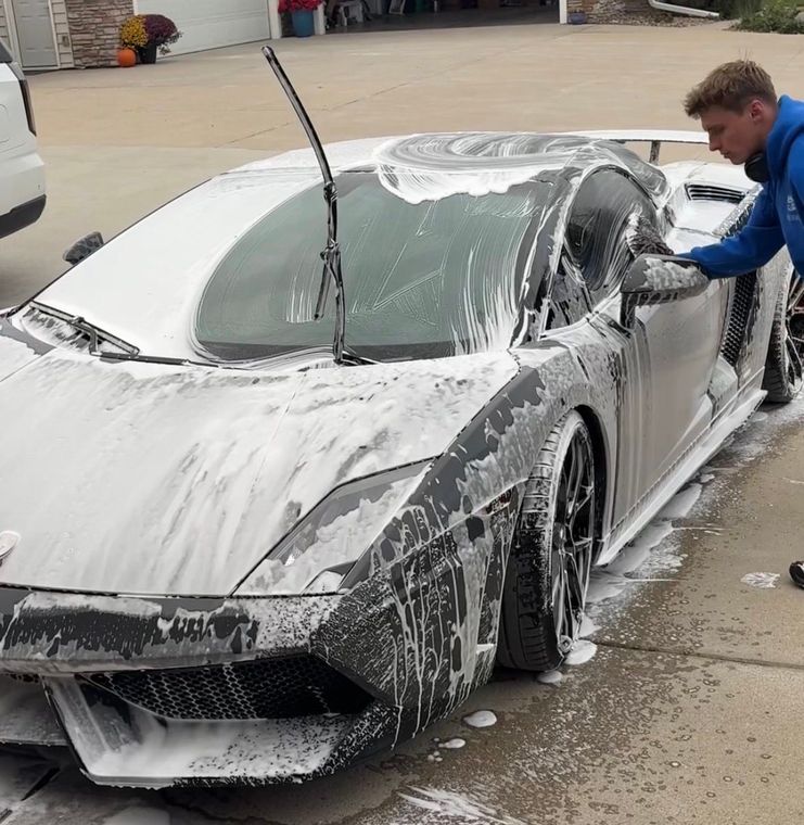 Person washing a white sports car covered in soap suds in a driveway