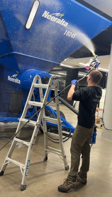 Person spray-painting a large blue boat hull while standing on ladders in a workshop