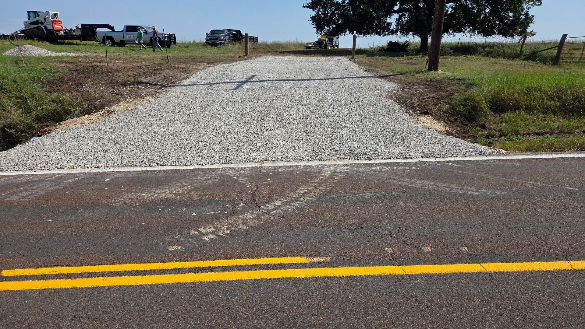 Gravel driveway entrance from asphalt road, vehicles and equipment in background, clear sky.