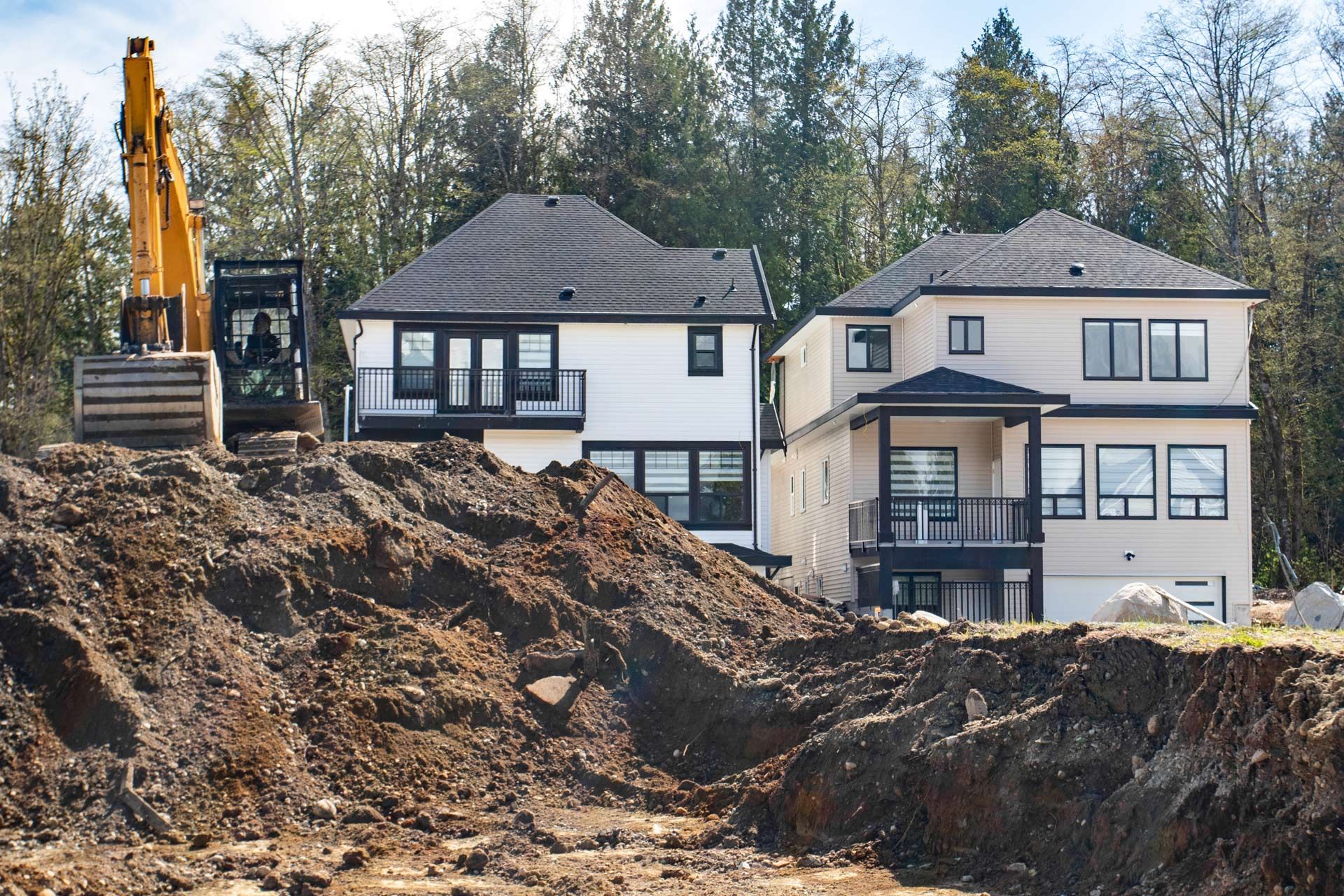 Excavator near two-story houses under construction, a pile of dirt in the foreground, and trees in the background.