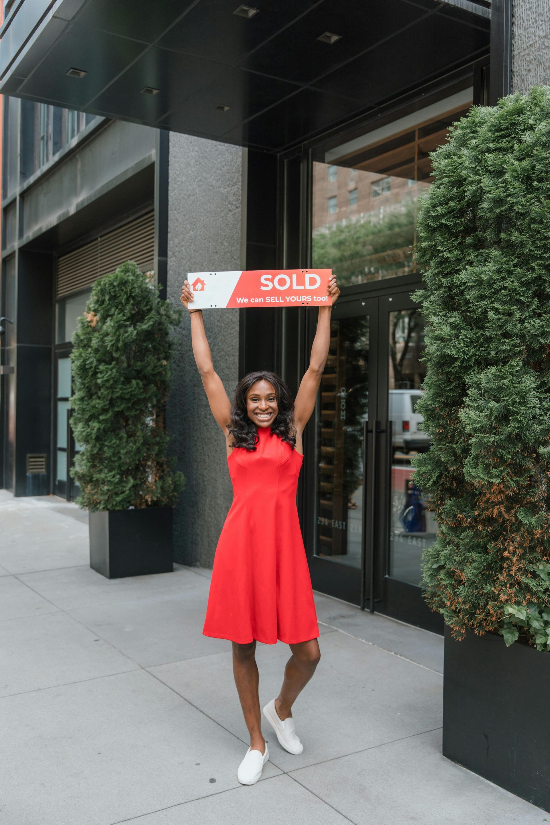 A woman in a red dress is holding a sold sign in front of a building.