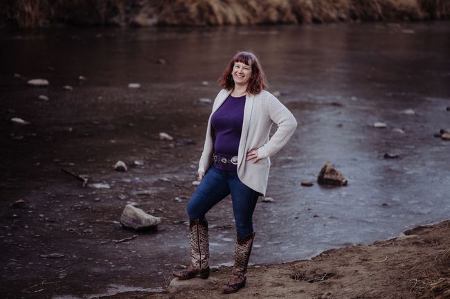 A woman in a purple shirt and cowboy boots is standing next to a body of water.