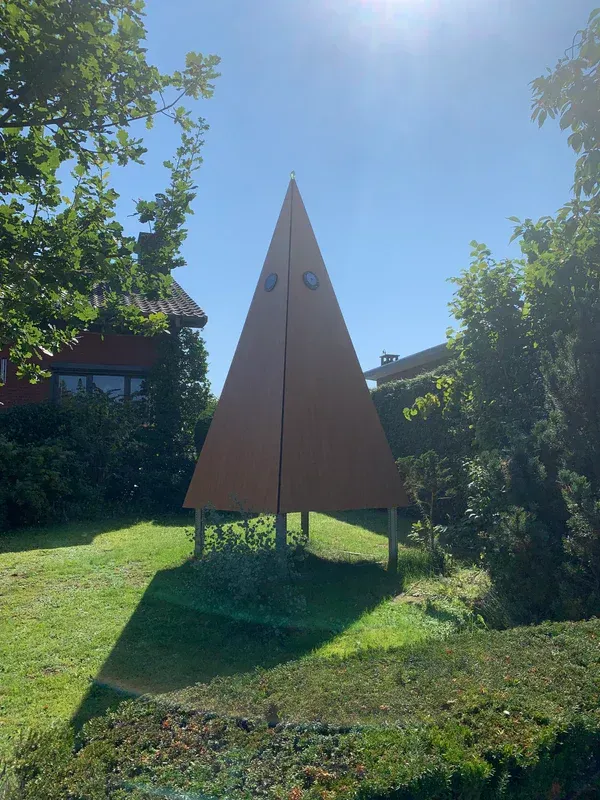 Een driehoekig beeldhouwwerk in een tuin met een huis op de achtergrond