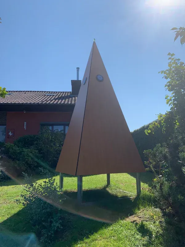 Core-piramide. Een grote houten piramide staat in het gras voor een huis.