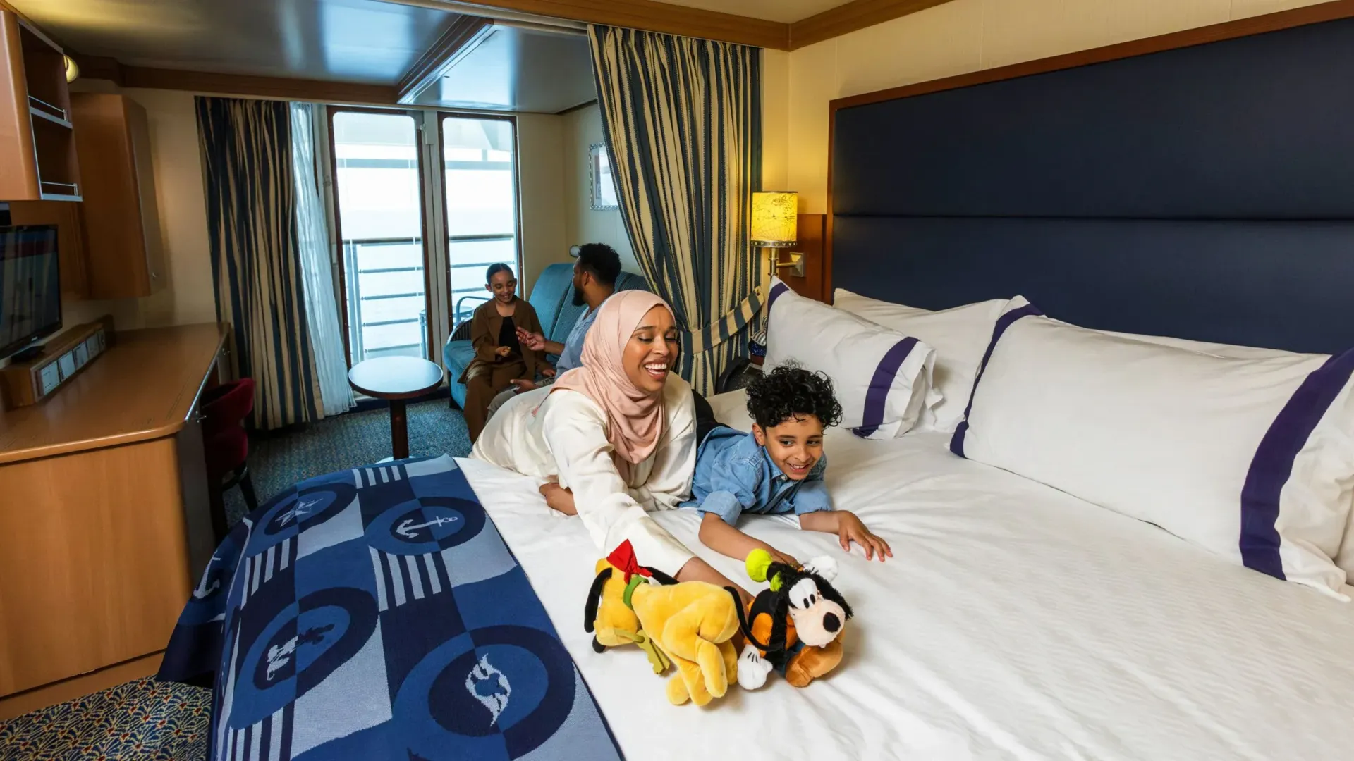Family on a cruise ship bed playing with plush toys, balcony visible in background.