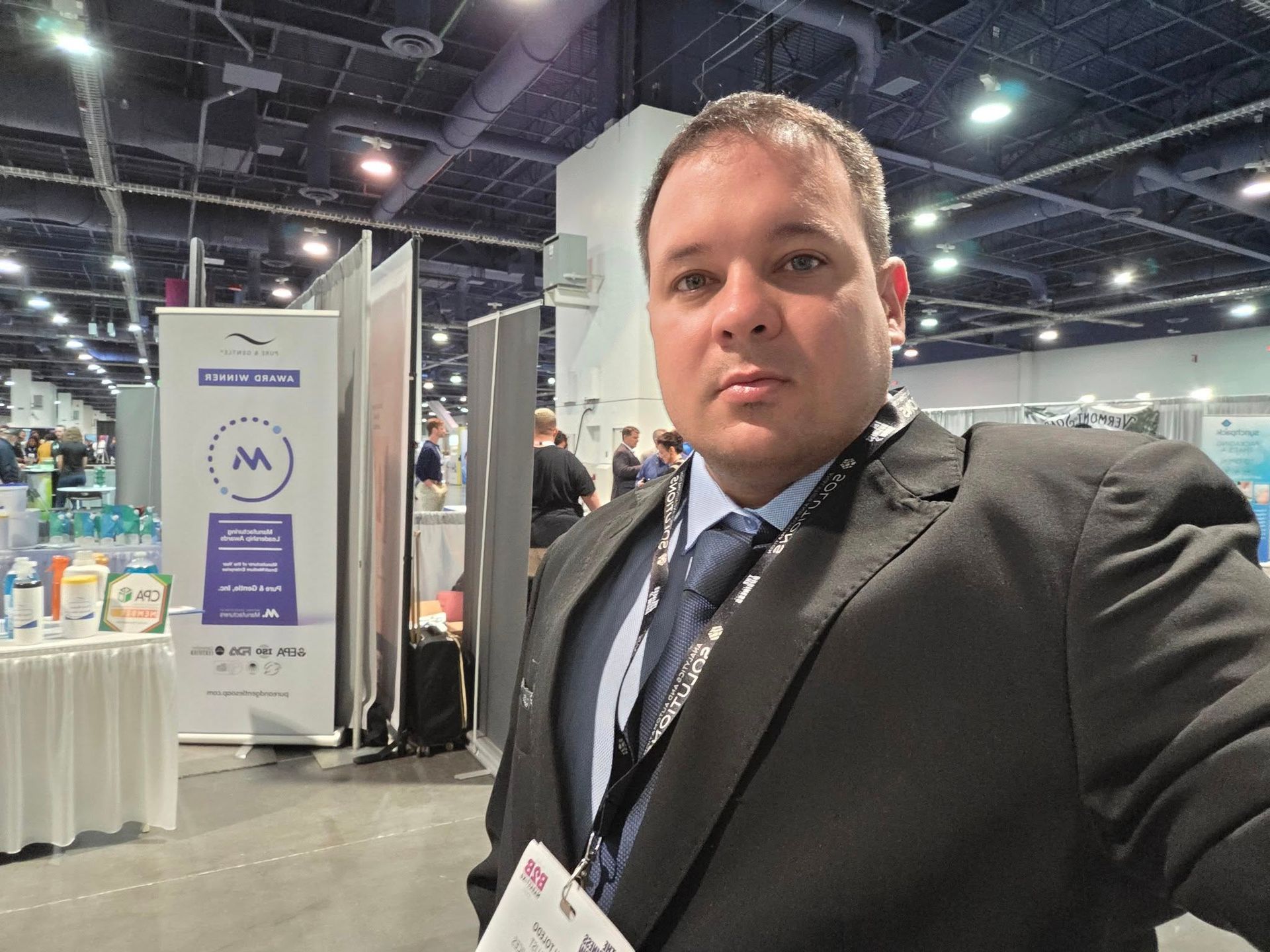 Man in a suit at a conference, looking at the camera. He is in front of a booth with banners.