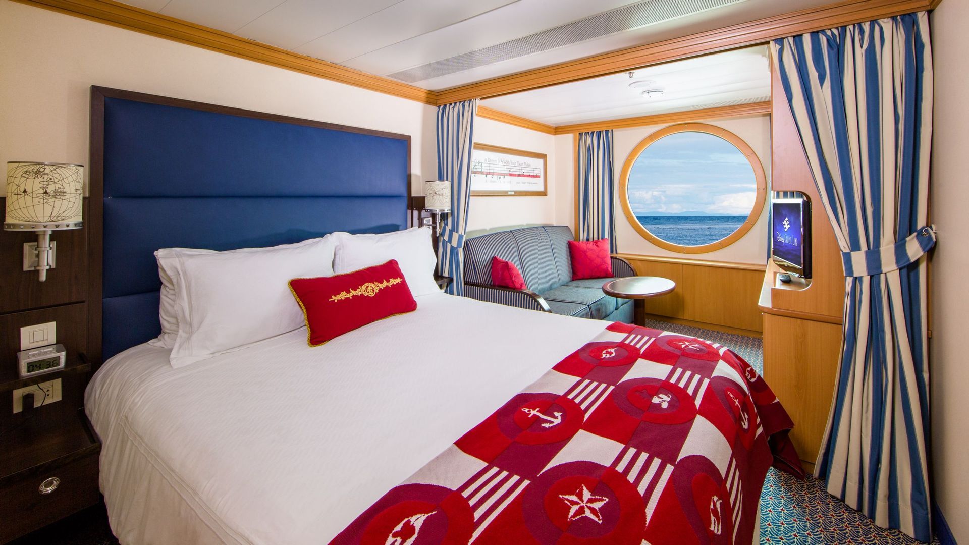 Cruise ship stateroom with a bed, porthole, sofa, and curtains; blue, red, and white nautical theme.