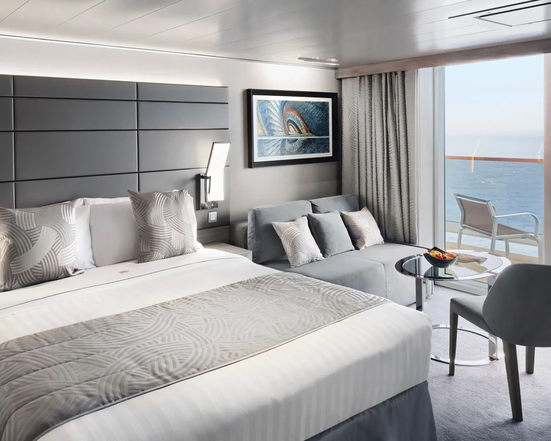 A bright cruise ship cabin with a bed, small sofa, and a balcony overlooking the sea.