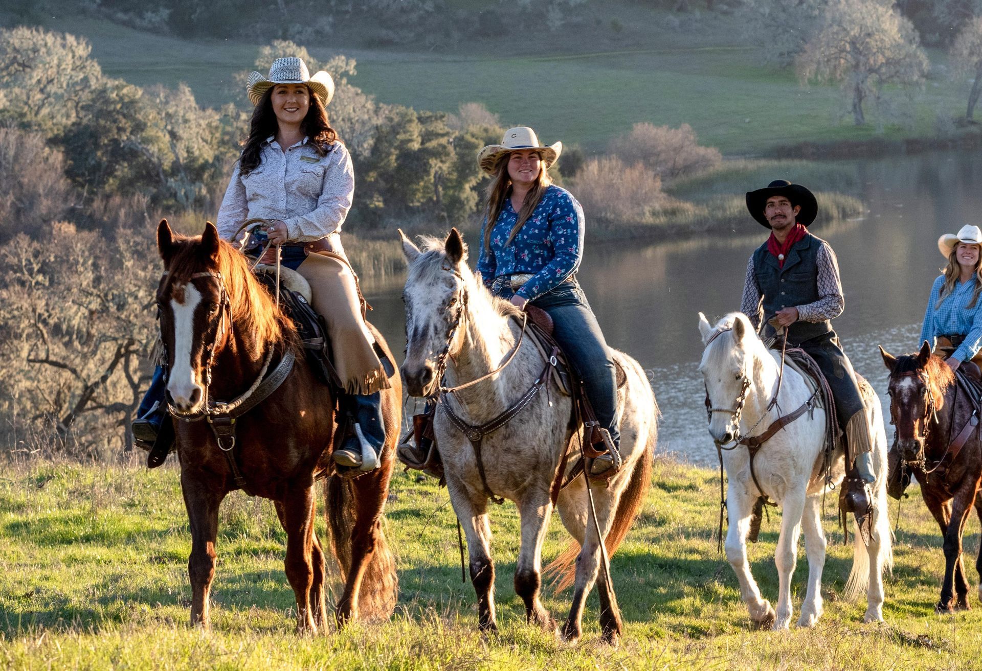 People horseback riding near a lake; riders wear cowboy hats and ride horses on a grassy hillside.
