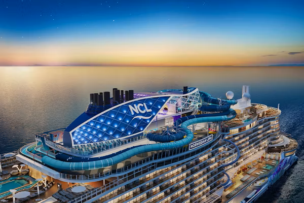 Aerial view of a Norwegian Cruise Line ship sailing on ocean at sunset; blue, white, and gold colors.