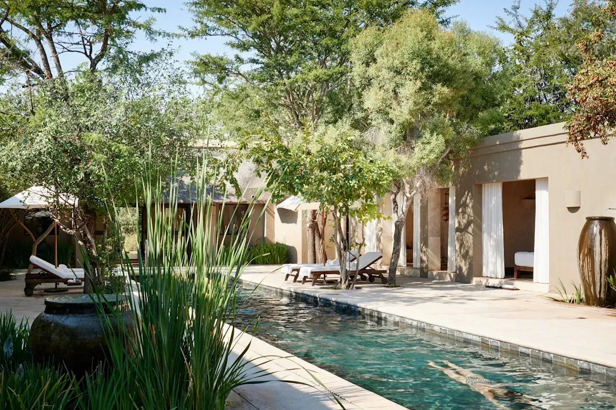Pool and lounge chairs at a luxury resort. Lush greenery surrounds the water, with beige buildings in the background.