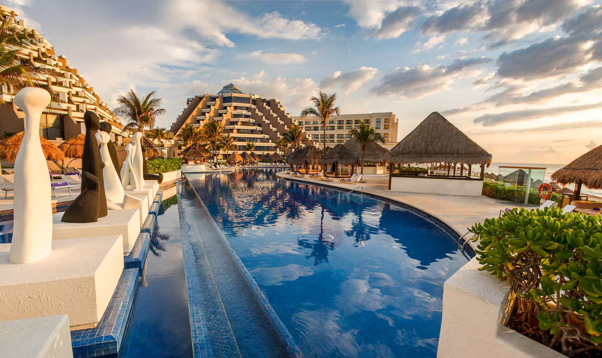 A resort pool curves toward a pyramid-shaped building, with palm trees and a thatched-roof structure.