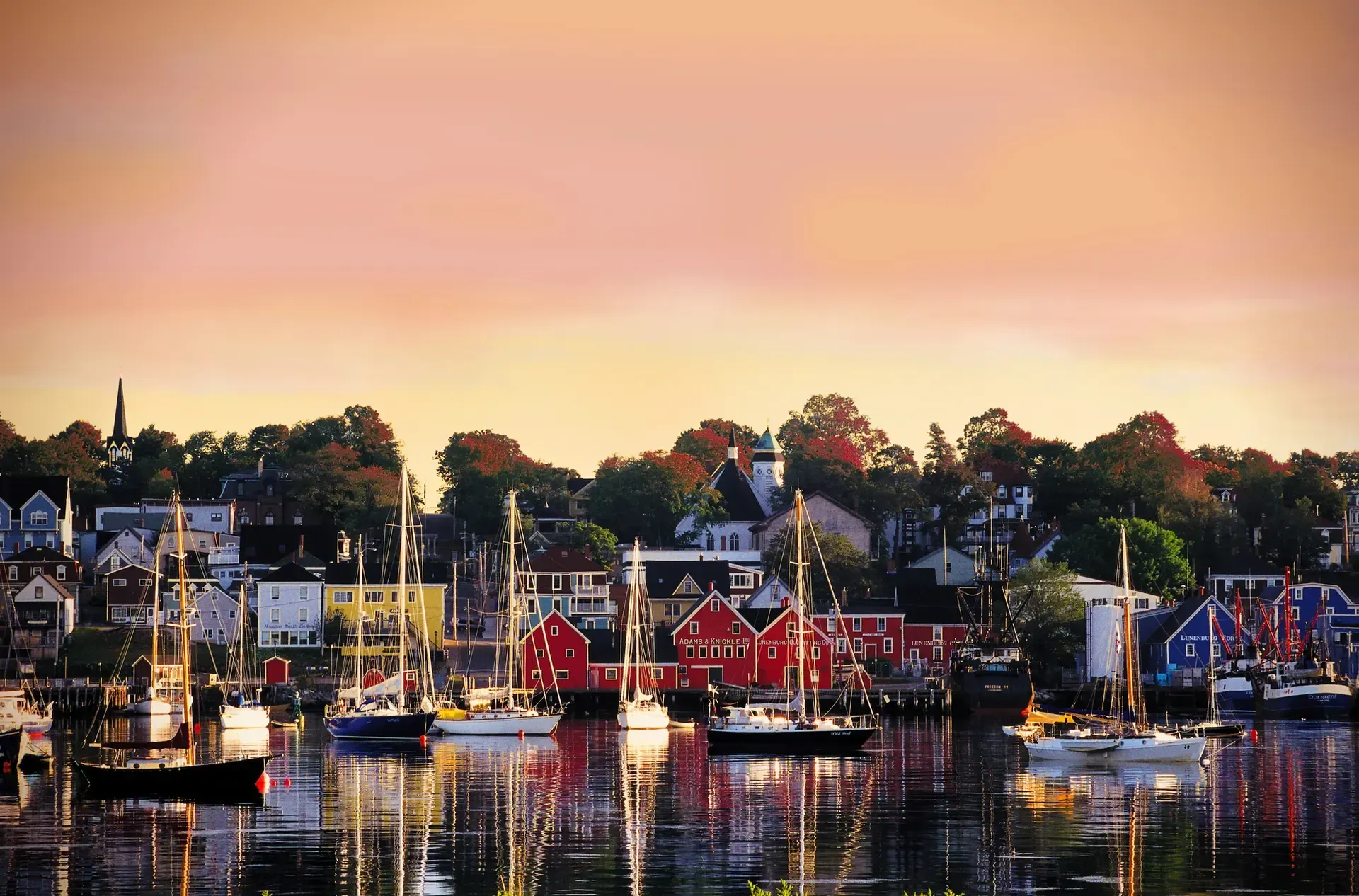 Boats on calm water in front of a colorful waterfront town under a pink and orange sky.