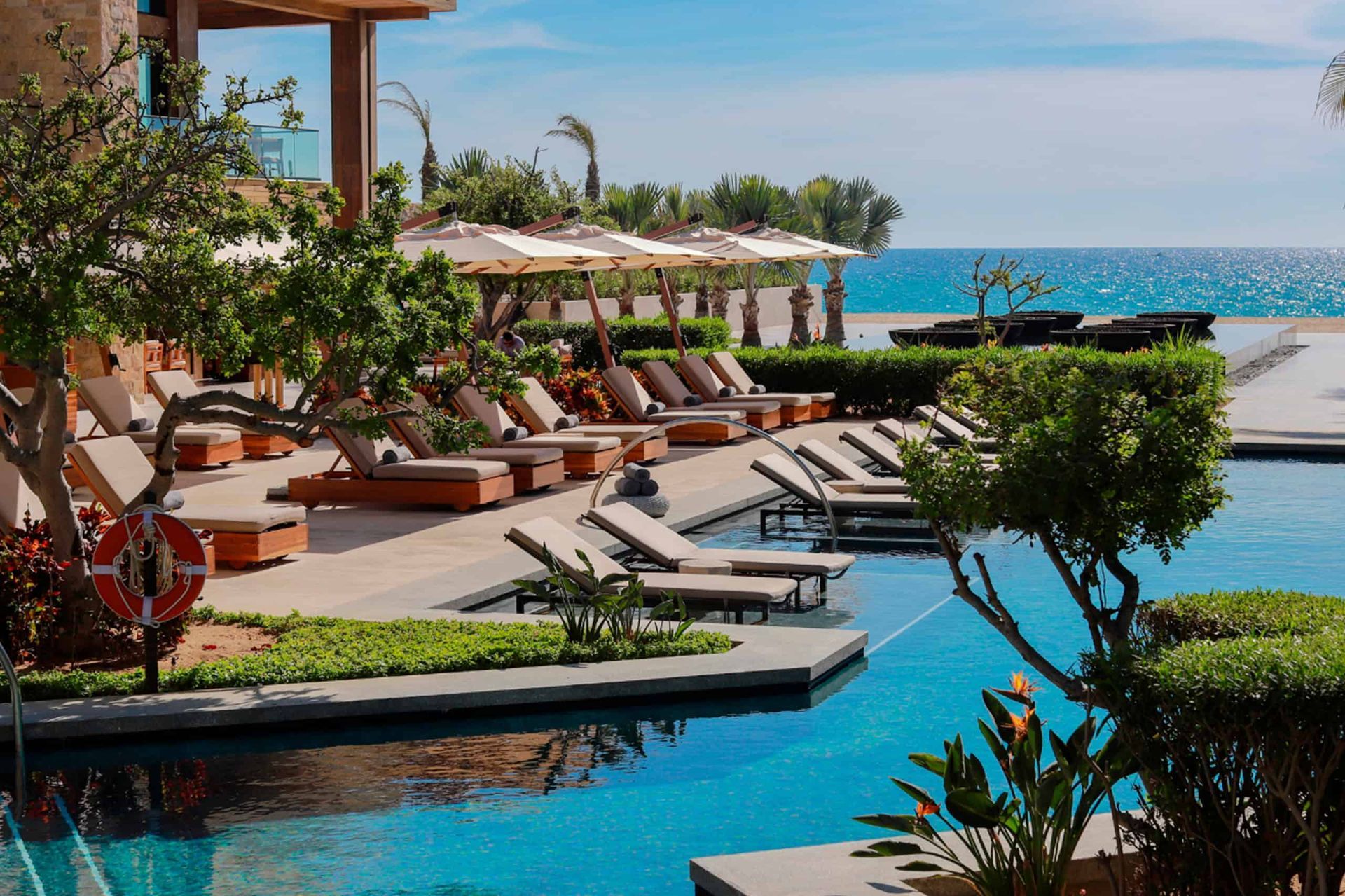 Poolside resort with ocean view; lounge chairs, umbrellas, trees, and blue water.