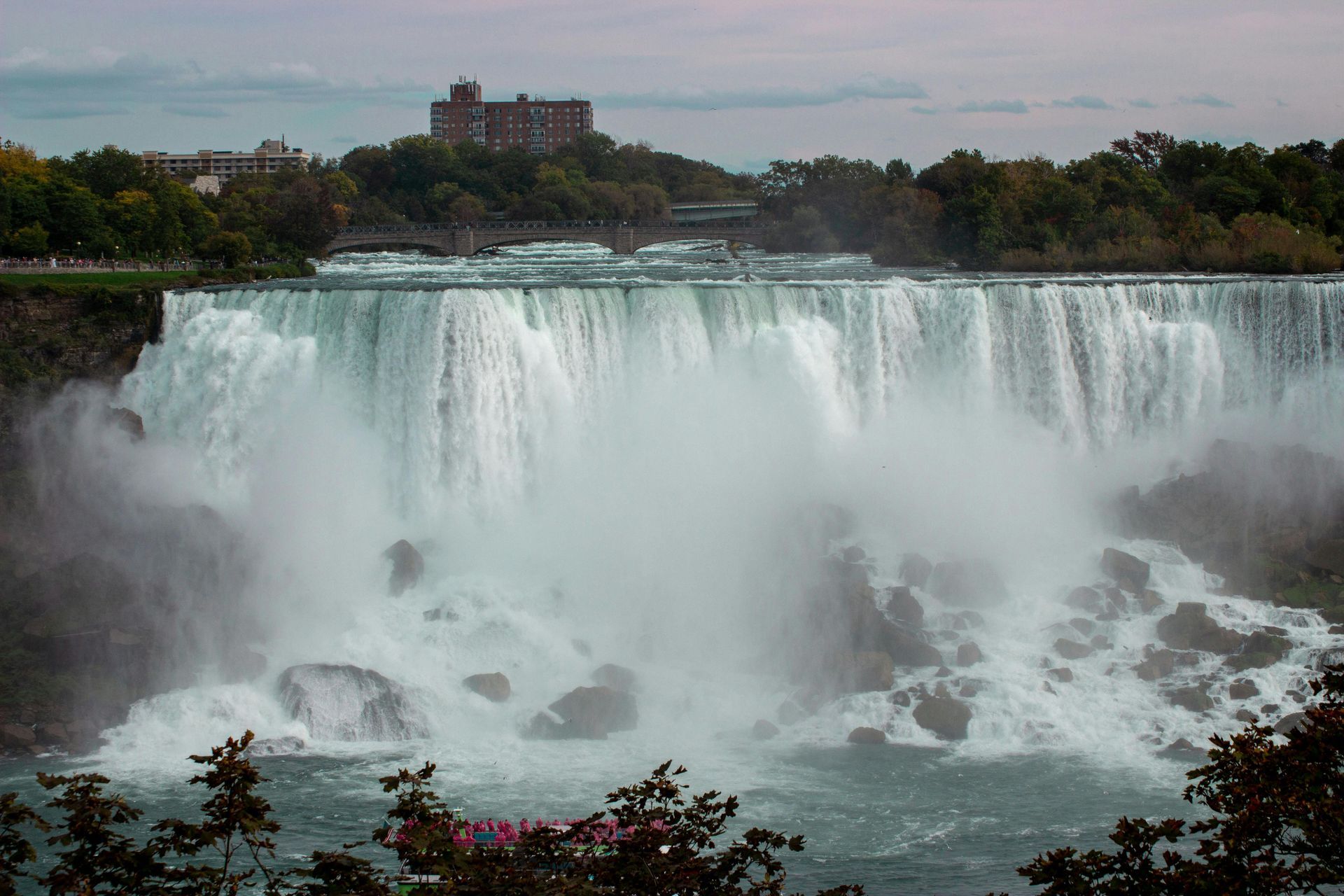Niagara Falls, cascading water. Buildings and trees on the horizon, overcast sky.
