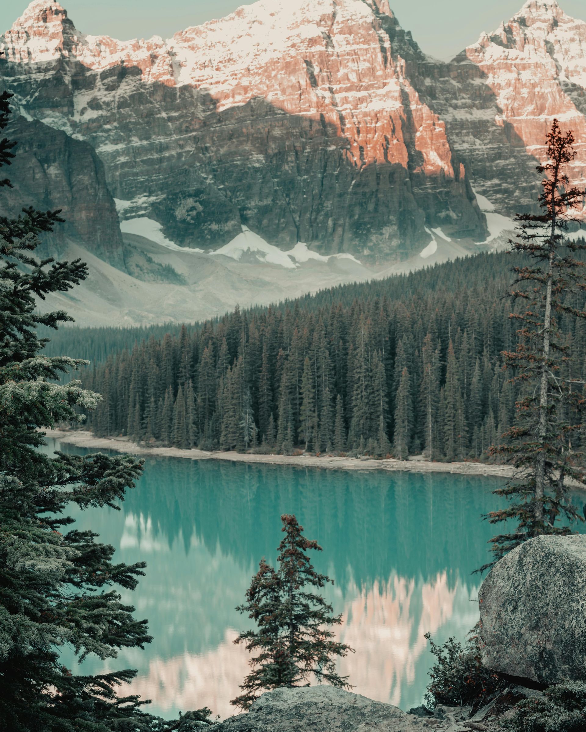 Snow-capped mountains reflect in a turquoise lake, surrounded by a dense evergreen forest.