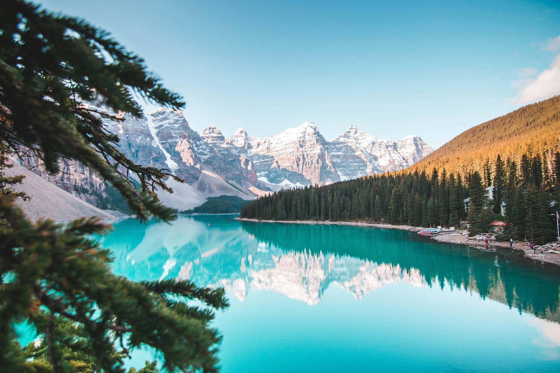 Turquoise lake reflecting snow-capped mountains and evergreen trees under a blue sky.