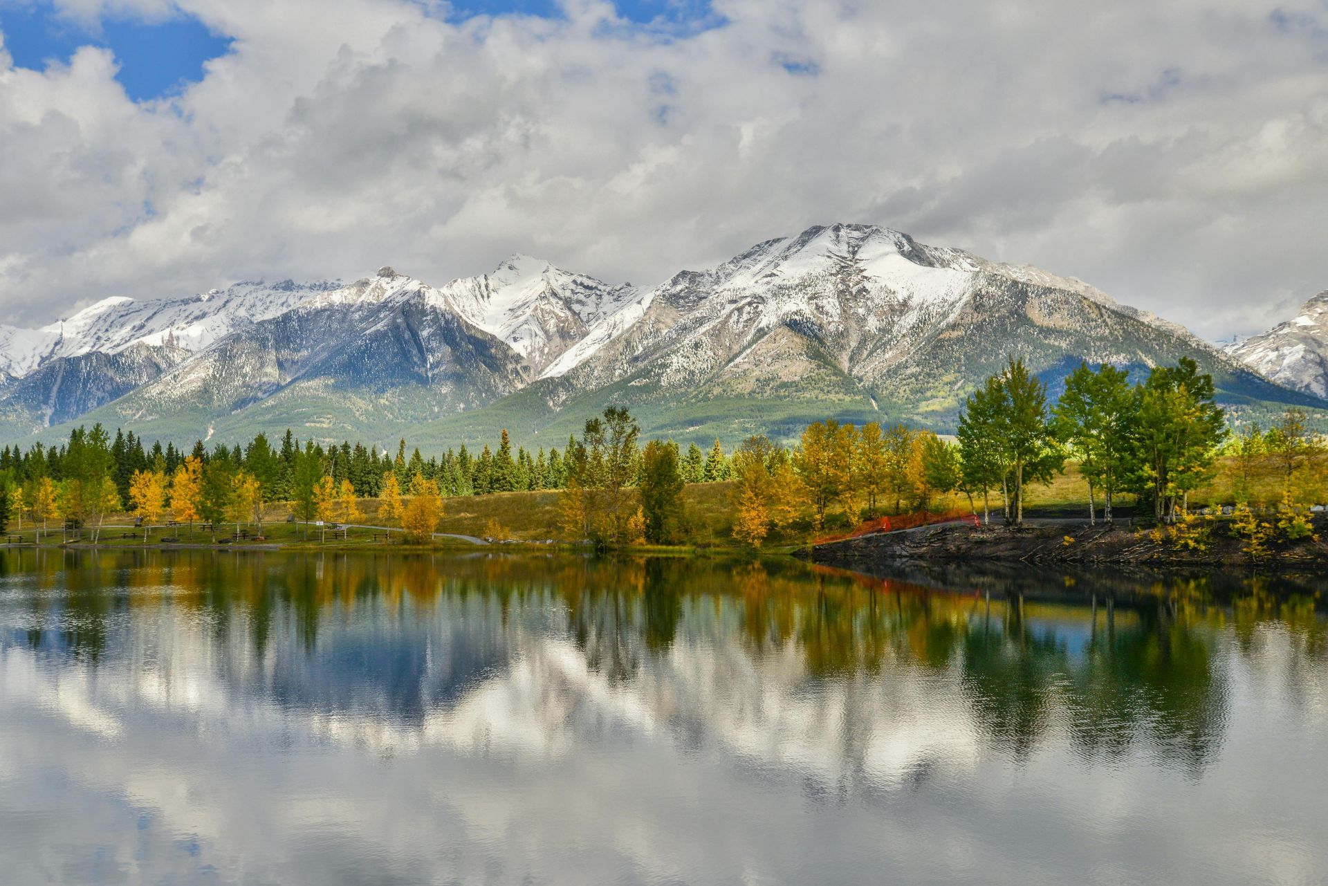 Snow-capped mountains reflected in calm water with fall foliage and partly cloudy sky.