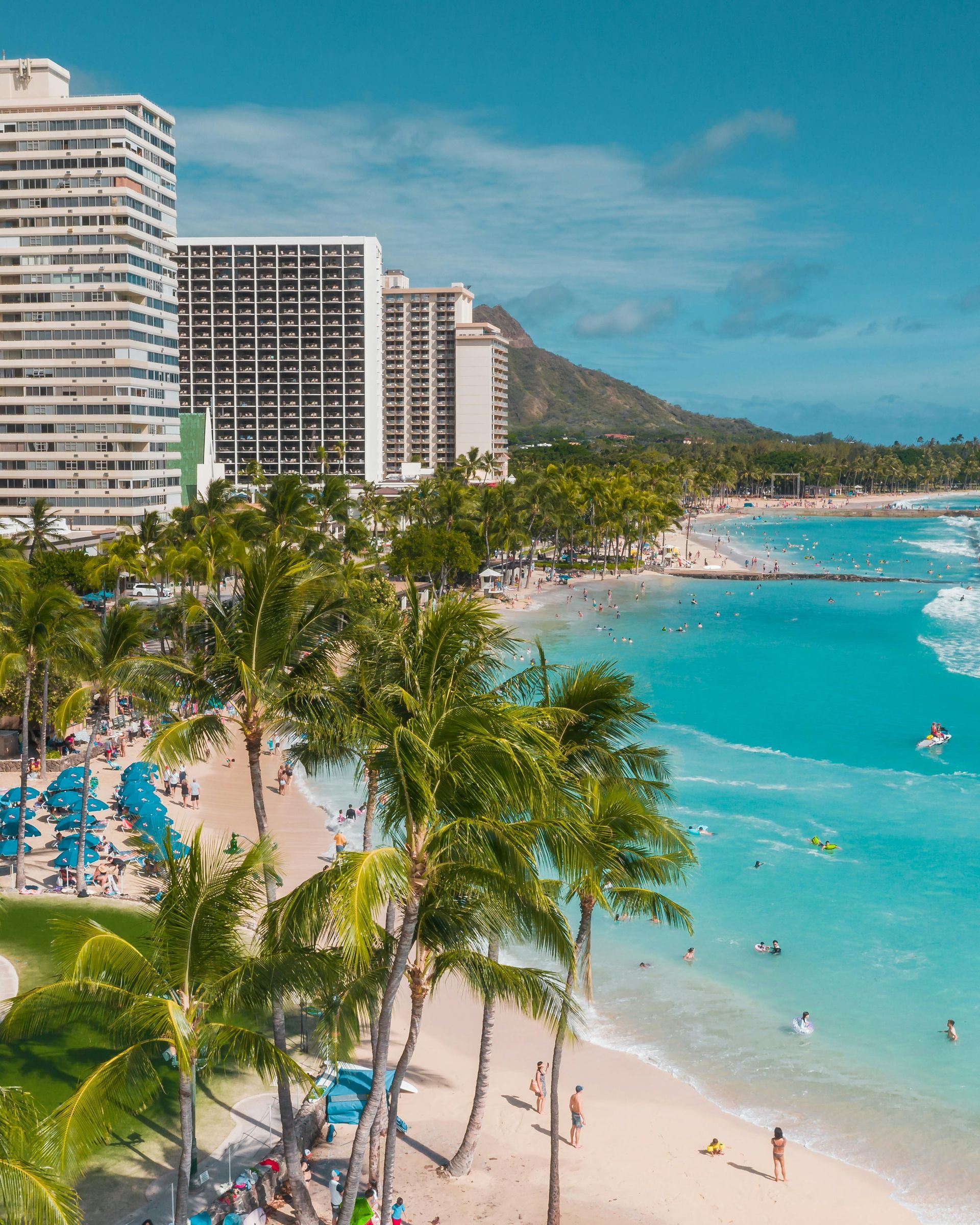 Beach with palm trees, turquoise water, buildings, and Diamond Head mountain in the background.