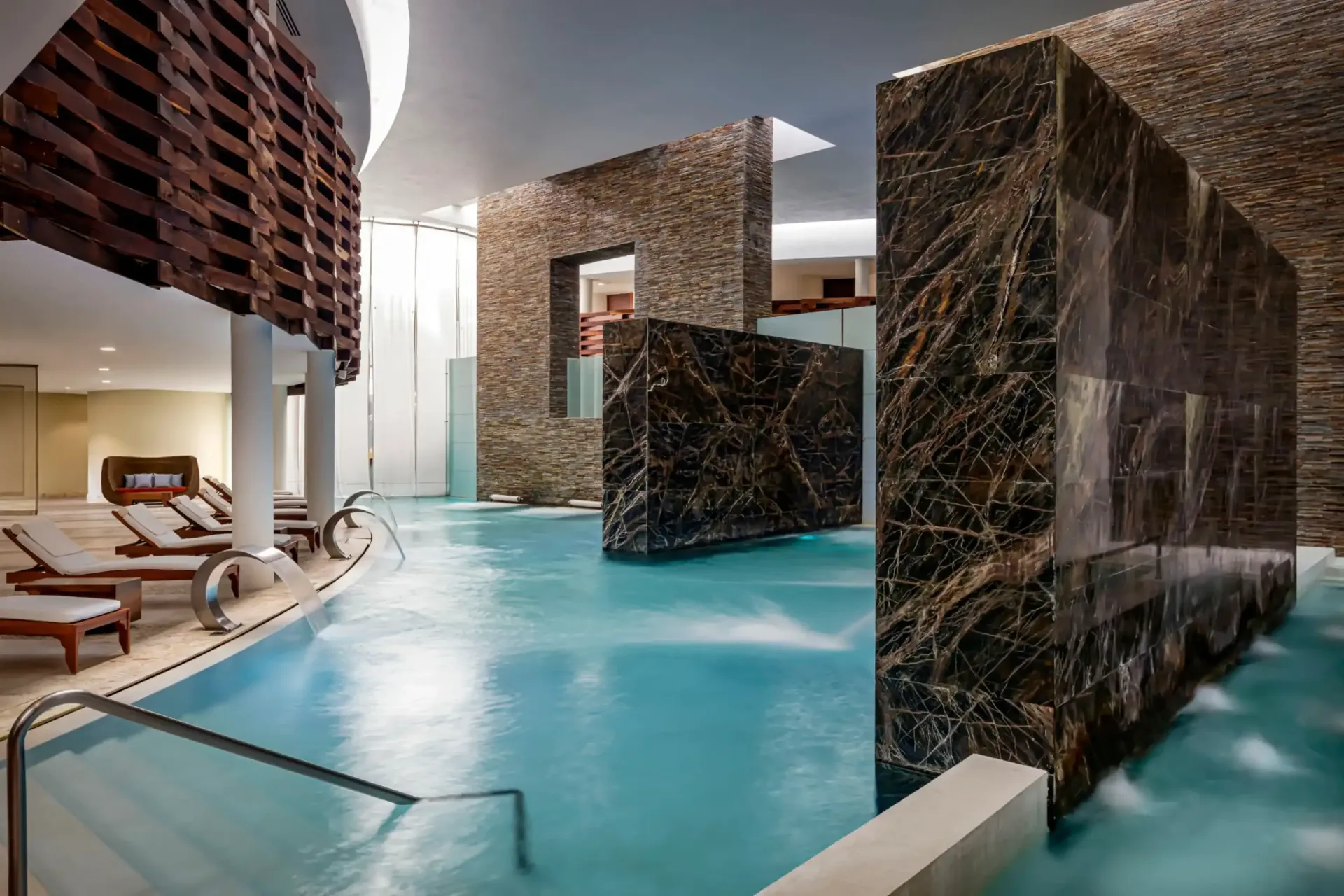 Indoor spa pool with blue water, stone walls, and lounge chairs.