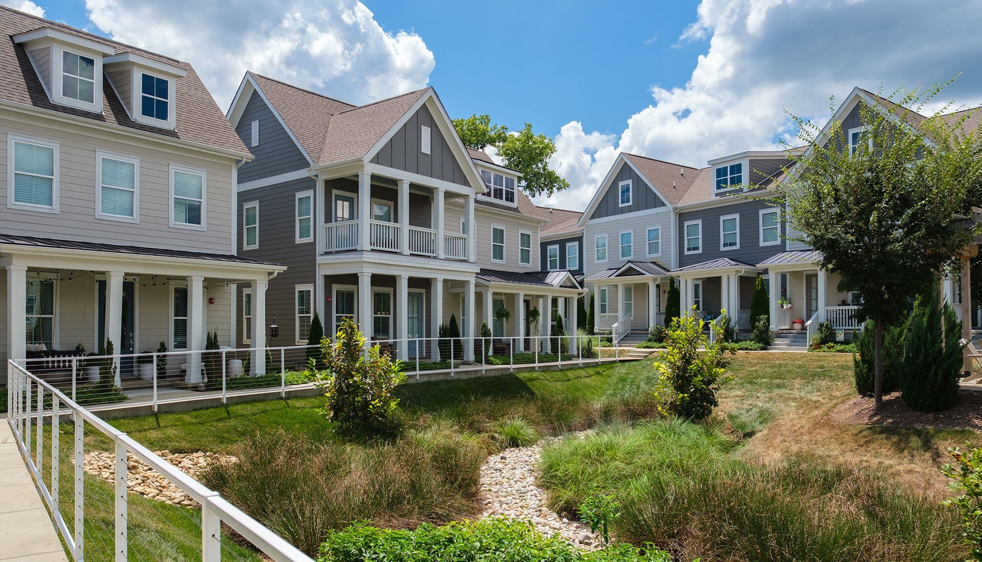 Row of townhouses, varying shades of gray and blue, with white trim and porch columns, fronting a grassy creek bed.