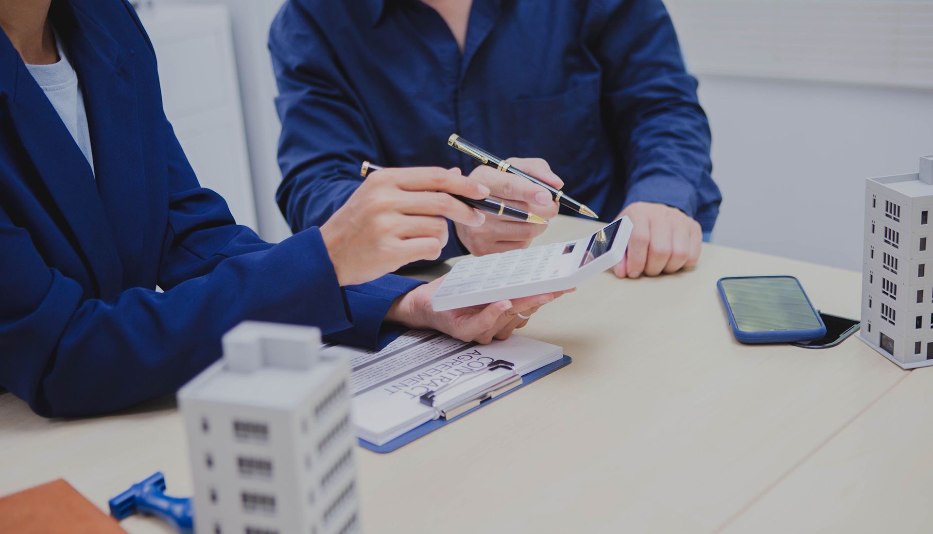 Two people reviewing building plans with calculator and miniature models on a desk.