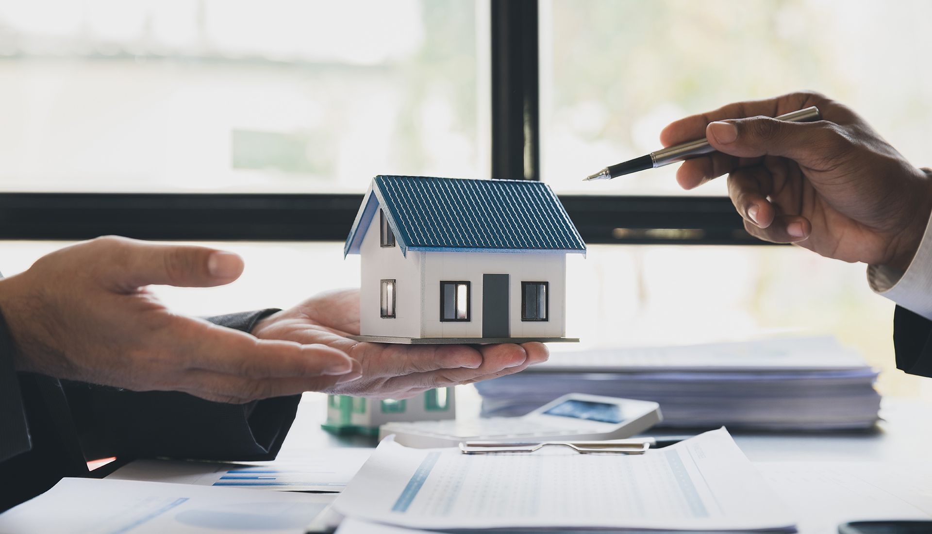 Two people exchanging a model house. One points pen at documents, others holds house.