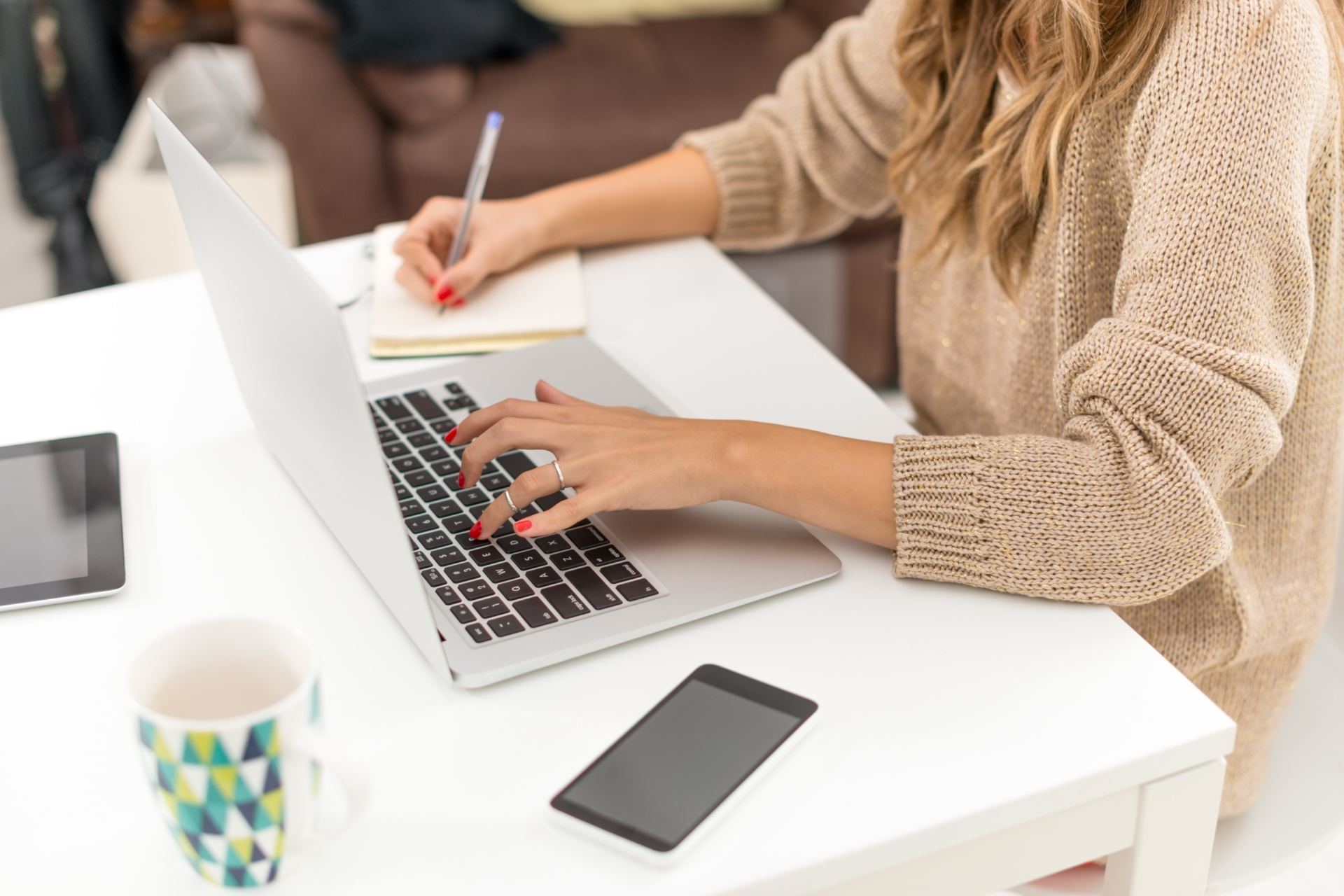 Woman working on a laptop and writing in a notebook at a white desk