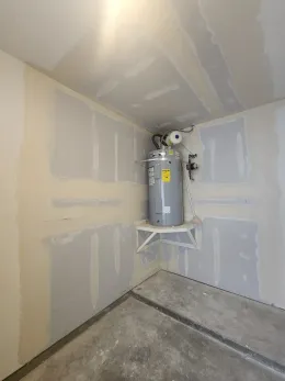 Water heater on a shelf in a corner, surrounded by unfinished drywall. Gray, white, and beige colors.
