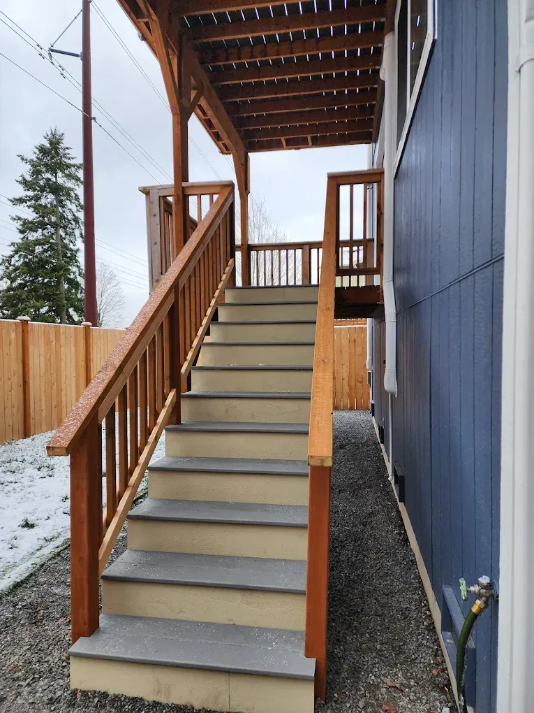 Wooden staircase leading to a covered deck, next to a blue building in a snowy yard.