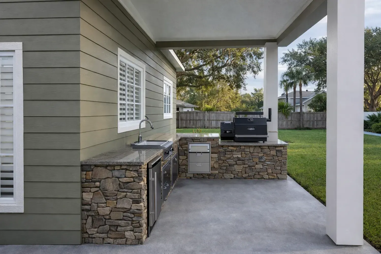 Outdoor kitchen with stone base, stainless steel grill, sink, and refrigerator under a covered patio next to a grassy yard.