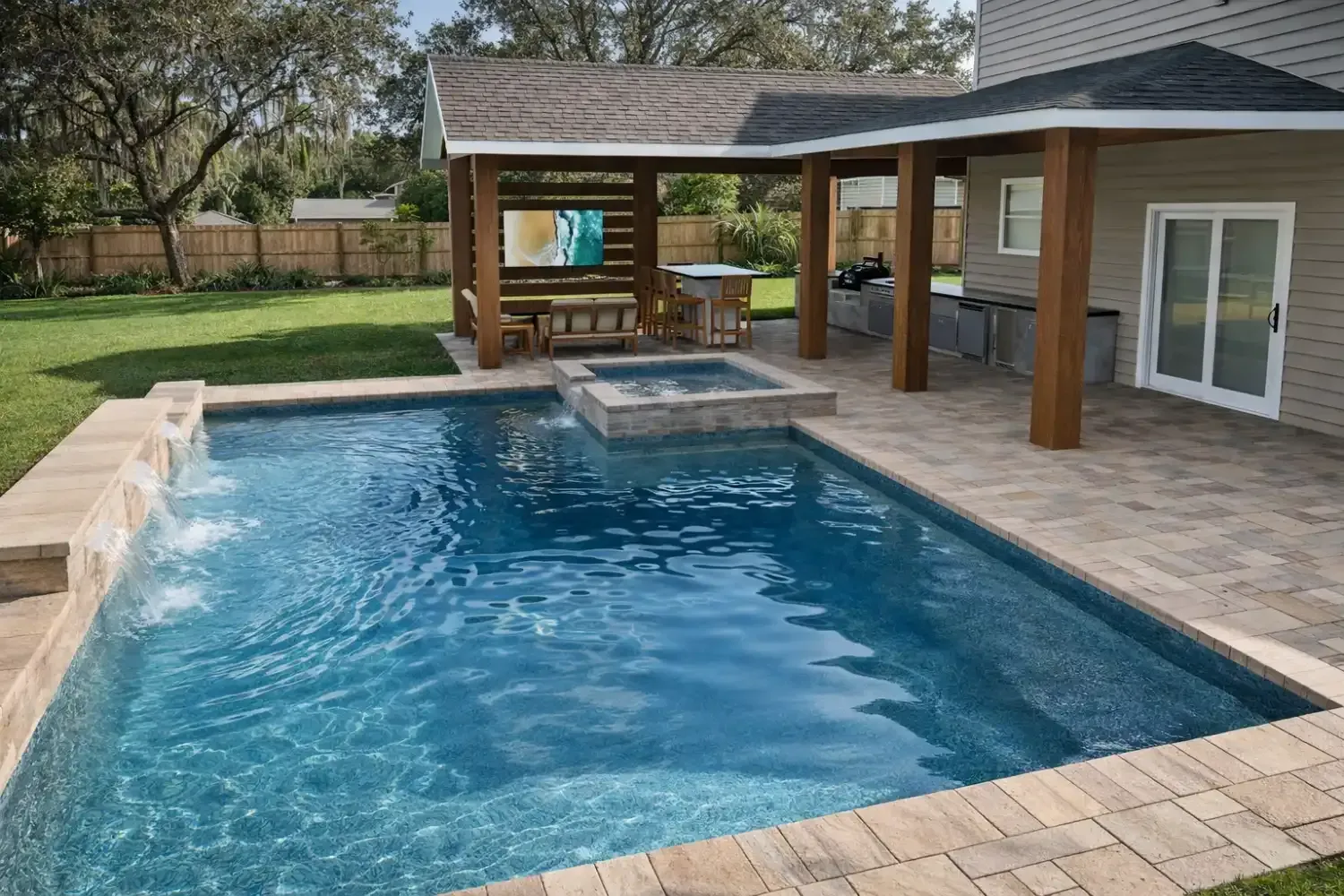 A blue backyard pool with a built-in spa and waterfall, featuring a covered patio area with pavers and a wooden structure.