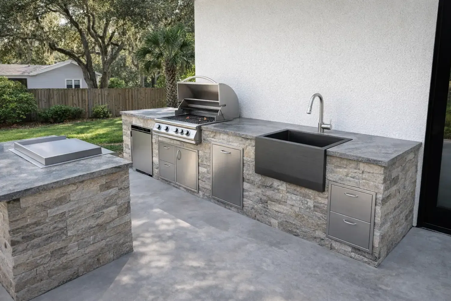Outdoor kitchen with a stainless steel grill, sink, and stone-veneer cabinets on a concrete patio.