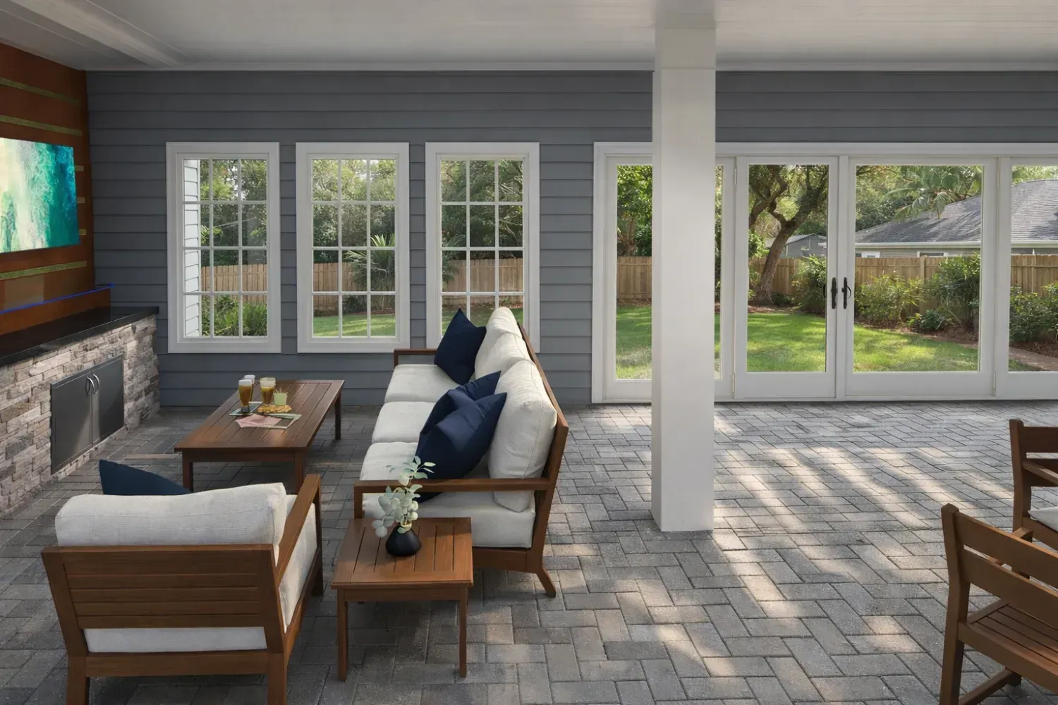 A bright covered patio with a stone fireplace, outdoor seating, and large windows looking out onto a green yard.
