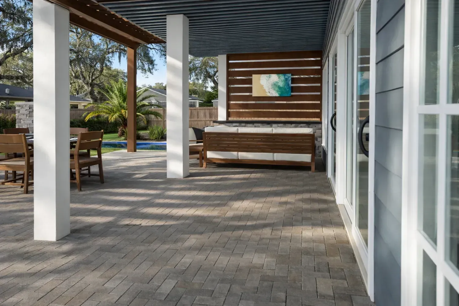 A covered patio with a wooden sofa, horizontal wood wall slats, and outdoor dining area on a paved ground surface.