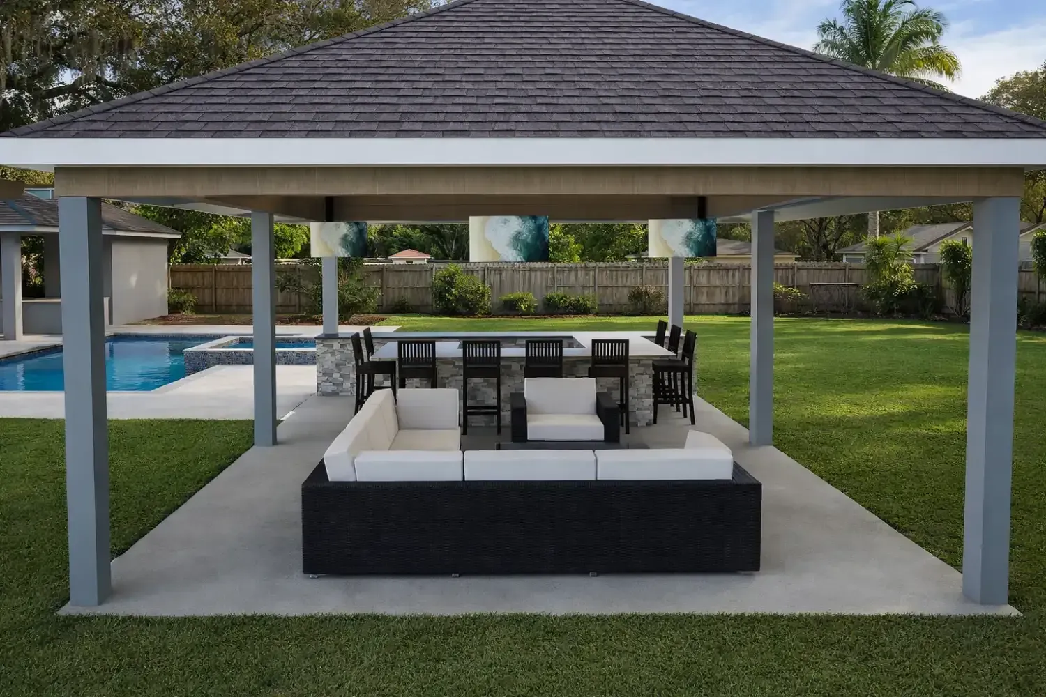 A covered backyard pavilion with an outdoor kitchen, bar seating, and a large sectional sofa facing a swimming pool.