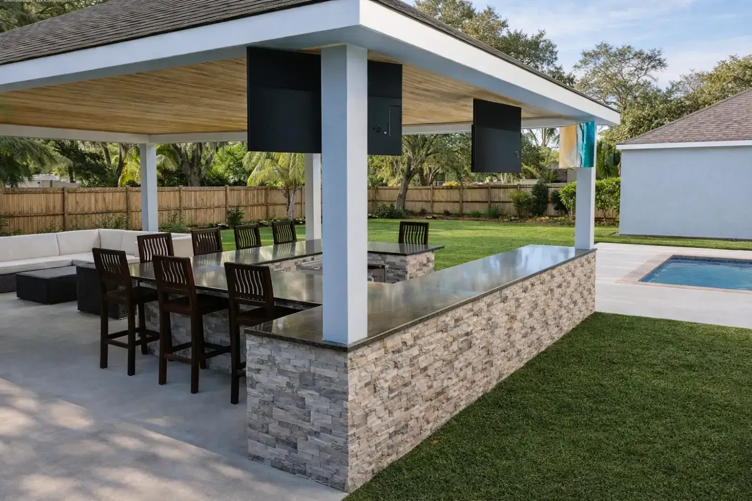 An outdoor covered kitchen and bar area featuring a stone counter, stools, and a patio overlooking a pool and lawn.