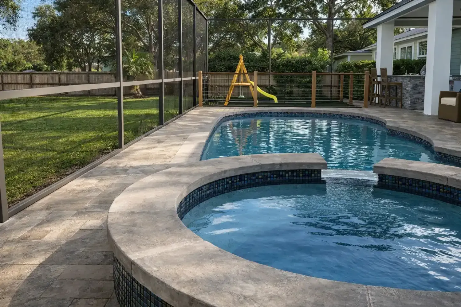 A backyard swimming pool and attached circular hot tub on a stone patio, enclosed by a screen, with a playset in the yard.
