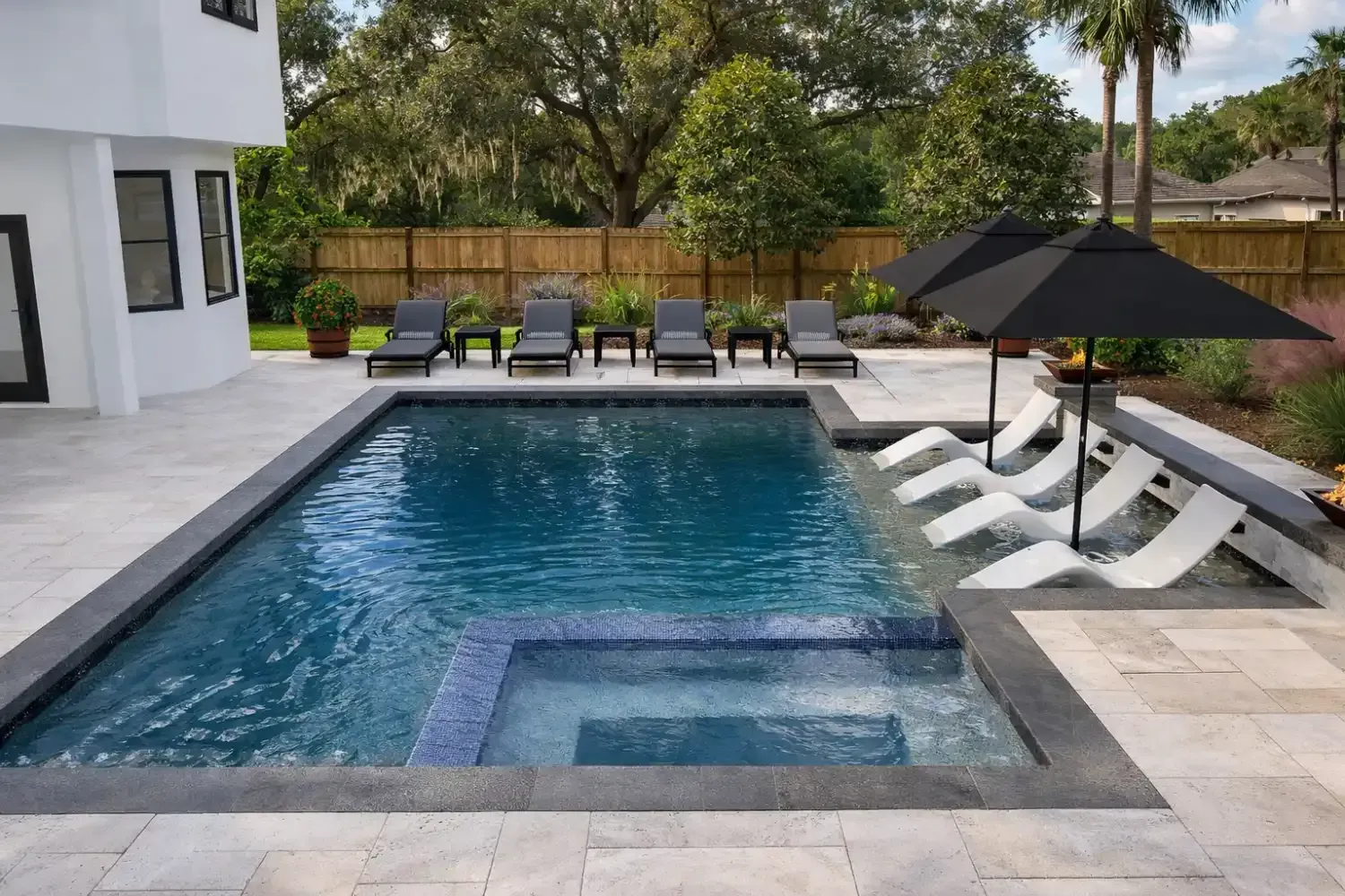 A backyard swimming pool with an attached spa, sun loungers, and patio umbrellas on a stone deck beside a white house.